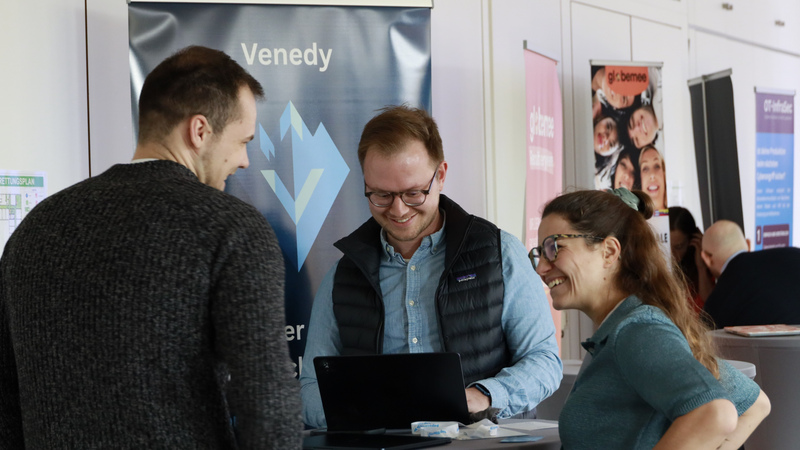 Networking-Szene beim B2B-DemoDay in der TechBase Regensburg. Das Start-up Venedy präsentiert seine Lösung auf einem Laptop. Ein anderer Teilnehmer hört interessiert und gut gelaunt zu. Ein dritter Teilnehmer (links) steht mit dem Rücken zur Kamera.