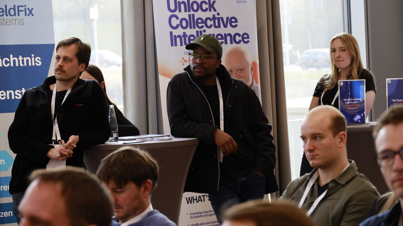 Blick ins Publikum während der Begrüßungsrede. Im Hintergrund stehen drei Personen an Stehtischen. Im Vordergrund sind einige sitzende Personen zu sehen, die ihren Blick nach vorne gerichtet haben.