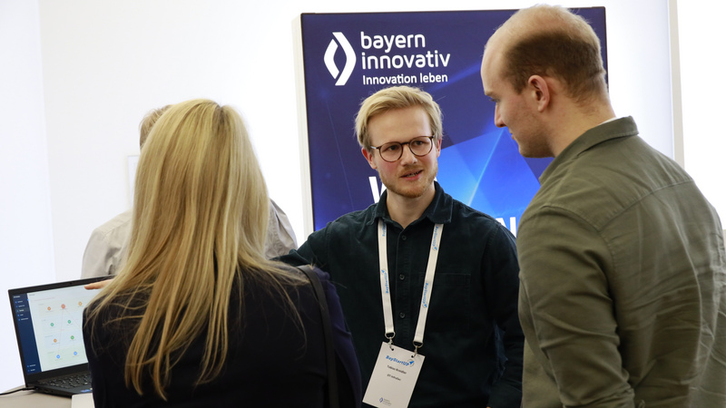 Drei Personen stehen gemeinsam am Stehtisch und schauen gemeinsam auf eine Präsentation, die auf einem Laptop läuft.